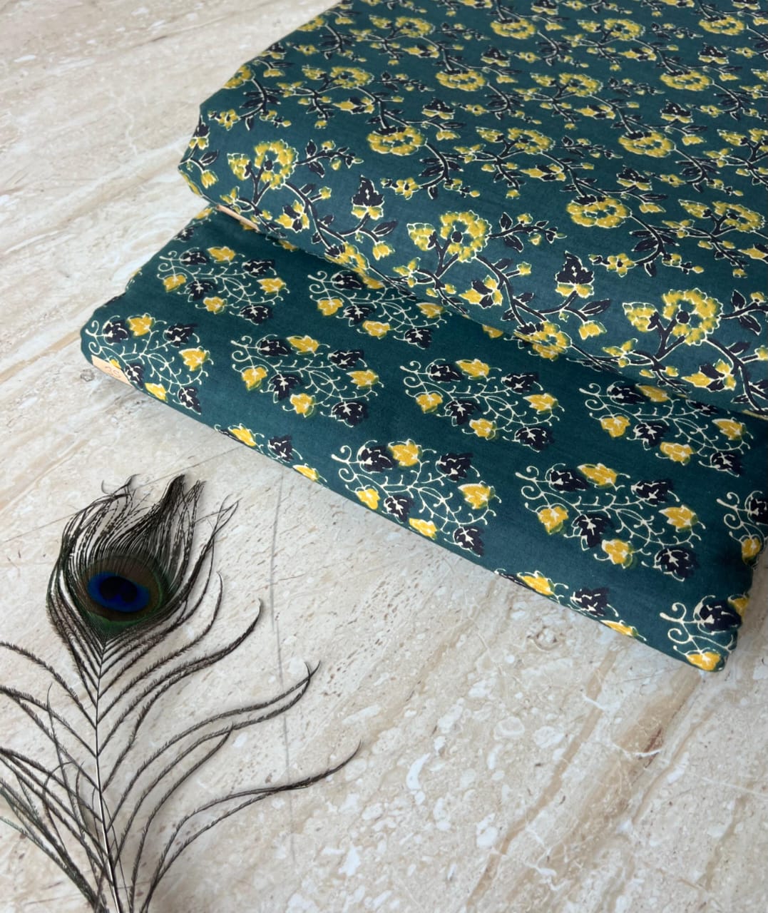 Two folded fabric sheets with a floral pattern on a light stone surface, accompanied by a peacock feather.