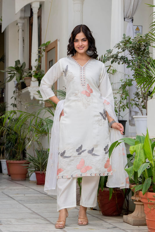 Woman in a white outfit with floral patterns standing in an outdoor setting with plants.